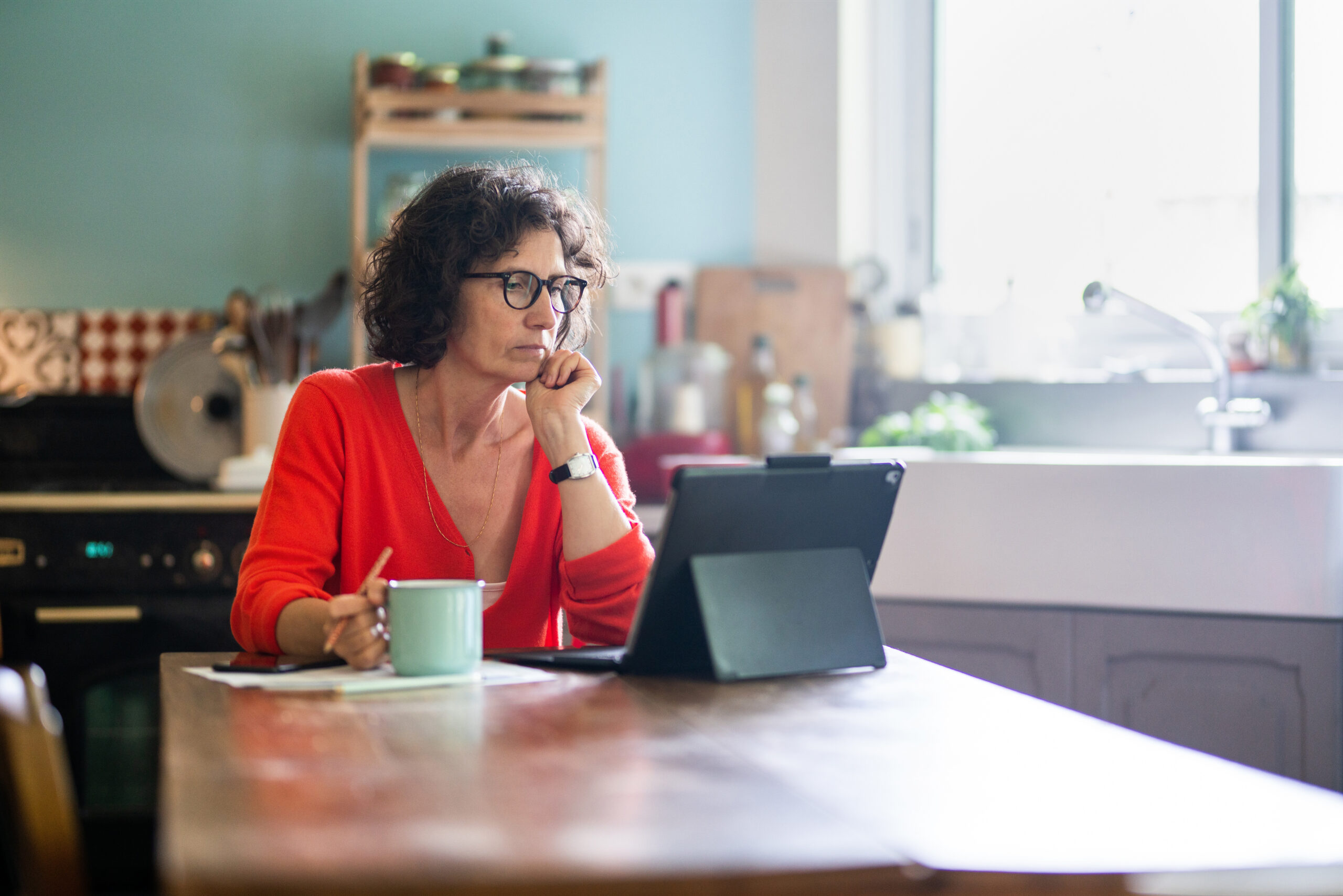 Beautiful middle-aged woman sitting in her kitchen, working on her laptop while drinking tea. 2022 Money Habits